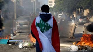 A Lebanese anti-government protester, wrapped in a national flag, stands in front of a road blocked with burning tyres and overturned rubbish bins (14 January 2020)
