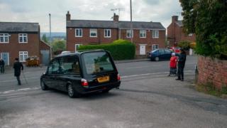 Hearse carries coffin as people watch