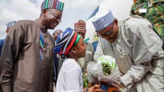 Buhari n gba ododo ikin kaabọ lọwọ ọmọdebinrin kan nigbati Gomina Orthom n wo o pẹlu ẹrin