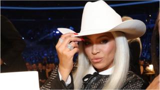 Beyoncé pictured at the 2024 Grammys wearing a white cowboy hat, pouts at the camera. The singer has bleached blonde hair worn loose and doffs her cap with her right hand, her nails painted bright red and chunky silver rings on her fingers. She wears a studded leather jacket and matching studded bow tie over a white shirt.