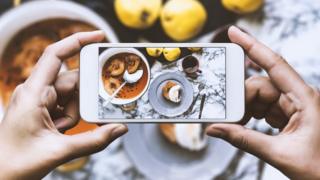 Hand taking top view photo of marble countertop. Cooking pot with poached quinces, poached quince with cream, fresh quinces and a cup of coffee on the countertop.