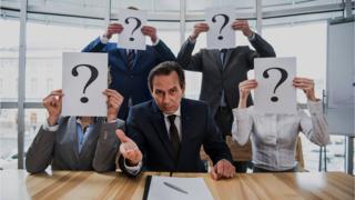 Personas tras un escritorio, con los rostros cubiertos con papeles con signos de interrogación