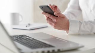 Stock image of a white male, face not pictured, on a mobile phone at his laptop