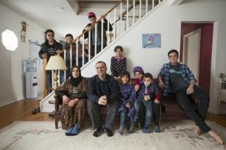 Изнутри семья Аль-Джасем представляет картину, начиная слева направо на диване, мать Савсан и отец Абдель Малек, их дети Валаа, Алаа, Хитам, Фадл и Рамаз, и стоящая на лестнице слева направо, Сулейман Абдель Малек , Маджид, Бачар и Ахмад в своем временном доме в Пиктоне, Онтарио, Канада,