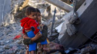 A Palestinian boy carrying a baby stands at a site of Israeli strikes in Rafah, Gaza