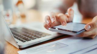 Una mujer frente a una computadora usando un celular
