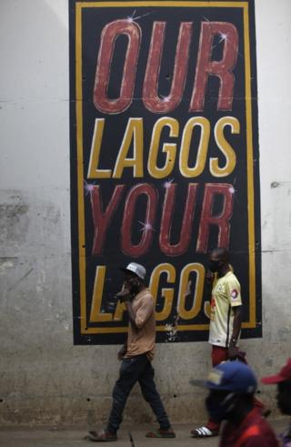 Men wearing face masks walk past a mural reading "Our Lagos, Your Lagos".