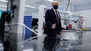 Trump de máscara em laboratório, fazendo sinal de joinha com o dedão