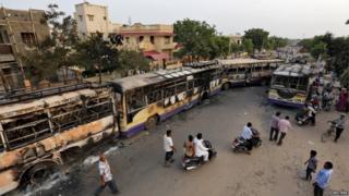 Пассажиры проезжают мимо поврежденных пассажирских автобусов, сожженных в результате столкновений между полицией и протестующими в Ахмедабаде, Индия, 26 августа 2015 года.