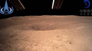 Foto mostra lado desconhecido da Lua. Na imagem, a Lua é vista com uma cor alaranjada. Há um pequeno buraco e, ao fundo, se vê o espaço, preto