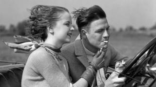 A young woman lights a cigarette for a young man driving a converible