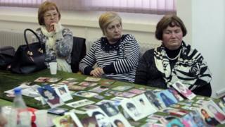 Фотографии жертв отображаются на столе, когда женщины смотрят, как Гаагский трибунал выносит свой вердикт по делу Караджича
