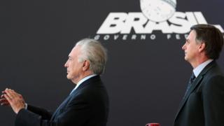 Michel Temer e Jair Bolsonaro olham para a mesma direção, com feição suave, durante evento em Brasília; Temer bate palmas