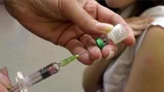 A generic photograph of a vaccine being drawn from a vial into a needle, while a person - most likely a child - bares their arm ready for the shot in soft focus in the background