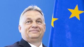 Виктор Орбан улыбается на голубом фоне, на котором виден флаг ЕС с ярко-желтыми звездами