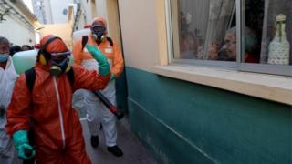 A worker wearing protective gear waves to a woman while using disinfectant to clean outside of a house in Santiago, Chile