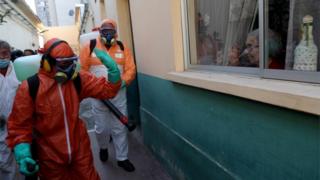 A worker wearing protective gear greets a woman as she uses disinfectant to clean outside a house in Santiago, Chile