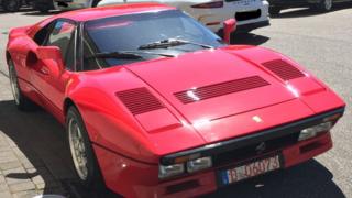 A Red Ferrari is seen parked against a curb in a parking area filled with other expensive vehicles - a pure white Porsche is seen behind it