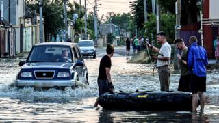 People stand next to an inflatable boat as a car drives past them in a flooded street of Kherson, Ukraine, 06 June 2023