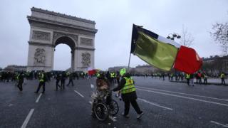 A demonstrator in a wheelchair is pushed by another, holding a French national flag with a yellow one attached to it, with the Arc de triomphe in the background during a Yellow Vests "Gilets Jaunes" anti-government demonstration in Paris on 12 January 2019