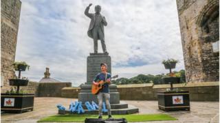 Cerddor yn canu dan gerflun Lloyd George ar faes Caernarfon