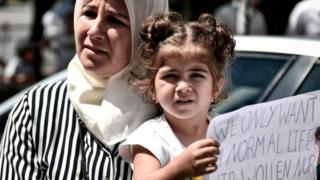 پناهجویان در مقابل سفارت آلمان در آتن؛ روی کاغذی که در دست این دختربچه پناهجوست نوشته شده: "ما فقط یک زندگی عادی میخواهیم"