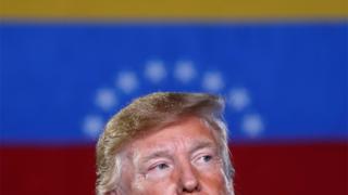 Donald Trump aparece olhando para o lado, com rosto pela metade, com bandeira da Venezuela ao fundo