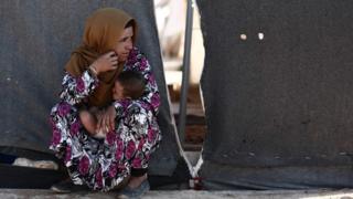Женщина несет своего ребенка, когда она сидит перед палаткой в ??лагере для перемещенных лиц в Аль-Гадфе, провинция Идлиб, Сирия (2 сентября 2018 года)