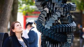 Женщина смотрит на скульптуру Token Homes Мэтью Пламмера Фернандеса