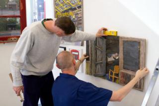 Two men hang artwork on a wall