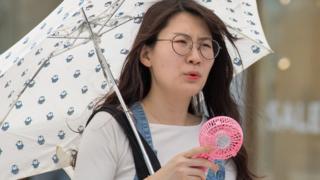A woman in Japan holds a pink fan to herself, carrying an umbrella to avoid heat