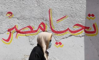 A woman wearing a protective face mask walks past a wall that has the words "Long live Egypt" painted in red and yellow.