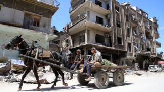 Two man sit on the back of a horse-drawn cart moving along a street, with ruined buildings stretching behind them