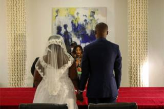 A couple dressed in wedding finery hold hands as the officiant stands before them.