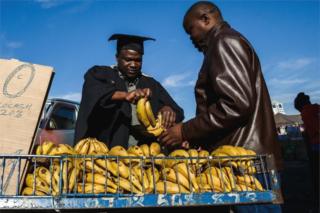 Abel Kapodogo (L), a graduate of Sociology from the University of Zimbabwe sells bananas along Robert Mugabe Road in Harare while wearing his graduation gown and cap in protest about the high unemployment levels in the country, July 20 2018.