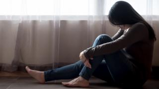 Imagem mostra mulher cabisbaixa, sentada no chão de uma casa