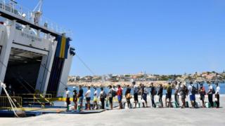 Migrants escorted by Guardia Di finanza police queue to board on a tourists ferry boat to Porto Empedocle in the Italian Pelagie Island of Lampedusa on July 29, 2020. - Boats with migrants mainly from Tunisia continue to arrive on the Italian island of Lampedusa. Intercepted by the boats of the Italian costal guard and guardia di finanza police before reaching the shore, migrants are escorted on the Island and transferred to the island's reception centre.