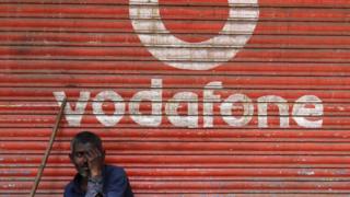 A man sits outside the downed shutters of a shop painted with a logo of Vodafone on its shutter in Mumbai, India on 24 February 2019