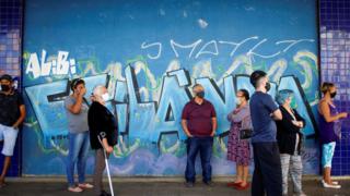 A queue of people wearing face masks in front of a wall covered in graffiti in Brasilia, Brazil