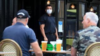 A doorman wearing PPE (personal protective equipment), of a face mask or covering as a precautionary measure against spreading COVID-19, stands on duty as customers sit with their drinks at a re-opened pub in Newcastle