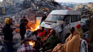 People sit around a fire next to rubble and damages near the site of a collapsed building in the aftermath of an earthquake, in Kahramanmaras, Turkey, February 8, 2023.