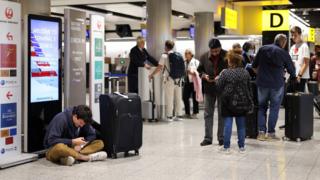 A man sits on the floor as others wait around in Heathrow Airport