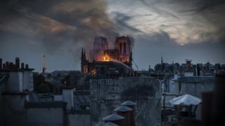 Foto mostra, no centro, catedral pegando fogo, em meio a prédios de Paris e com Torre Eifel ao fundo durante anoitecer
