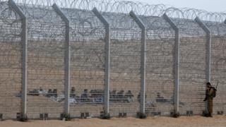 An Israeli soldier stands near the border fence between Israel and Egypt as African would-be immigrants sit on the other side near the Israeli village of Be'er Milcha, in this September 6, 2012 file photo