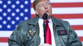Donald Trump fala no microfone com bandeira dos EUA atrás