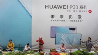 Women sell vegetables in front of a billboard advertising smartphones for China's Huawei Technologies Co., on June 1, 2019 in Mangshi, Yunnan Province, southwestern China.