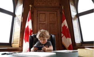 Трюдо младший сидит за столом премьер-министра и играет со смартфоном в окружении двух канадских флагов