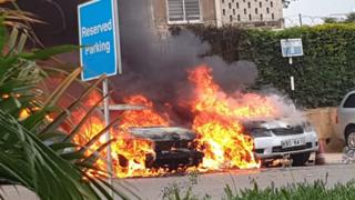   In the parking lot around the complex, cars are burning around him 