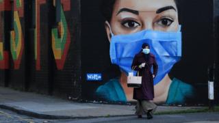 Una mujer con una máscara camina frente a un mural de un trabajador del NHS