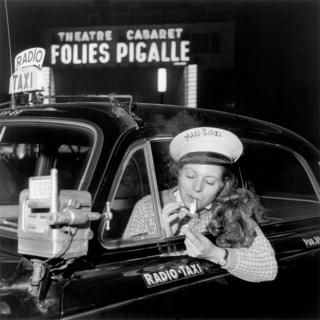 A woman sits in a taxi driver seat and lights a cigarette